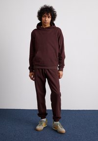 Felpa bordeaux con cappuccio e pantaloni coordinati in morbido tessuto. Presenta una tasca frontale, cappuccio con lacci e polsini elasticizzati. Carnagione con capelli ricci.