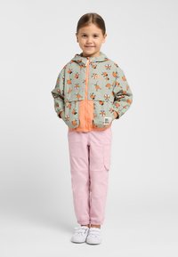 Veste coupe-vent vert clair avec des accents orange, arbore un motif de dessin animé, des poches avant et des poignets élastiques. Associée à un pantalon cargo rose.