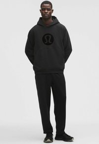 Fekete kapucnis pulóver nagy fekete logóval az elején, fekete nadrággal párosítva. Puha pamut keverékből készült, elülső zsebbel rendelkezik.