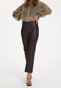 Genomskinlig blus med leopardmönster i brunt och ballongärmar, som matchas med högmidjade, bruna byxor i läderliknande material. Svarta sandaler med remmar fullbordar looken.