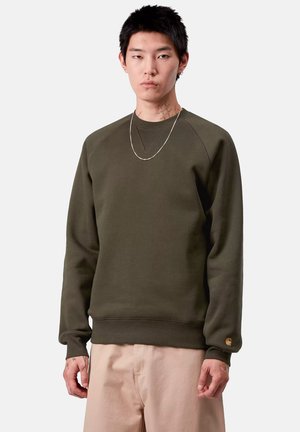 Joven con cabello corto y negro que lleva un sudadera verde oliva, pantalones beiges, una cadena de plata y tatuajes visibles en el cuello y las manos.