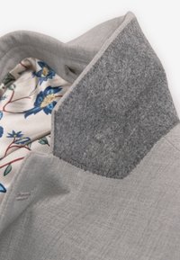 Giacca grigia con collo in feltro dalla texture. Interno con tessuto decorato da motivi floreali blu e beige. Vista ravvicinata dei dettagli.