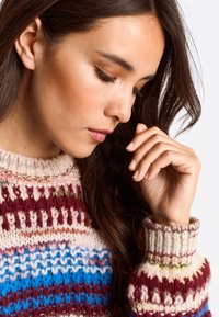 Megztinis su daugiakoloriu raštu, apimančiu raudoną, mėlyną ir kreminę spalvas; raukšlėtas apykaklė ir rankogaliai; švelni tekstūra ir masyvi forma.