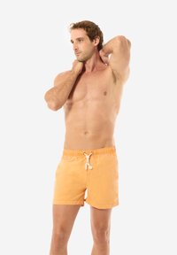 Shorts da bagno arancioni realizzati in un tessuto liscio, con vita regolabile tramite cordino e fessure laterali per una maggiore libertà di movimento. Il modello posa con le mani dietro il collo.