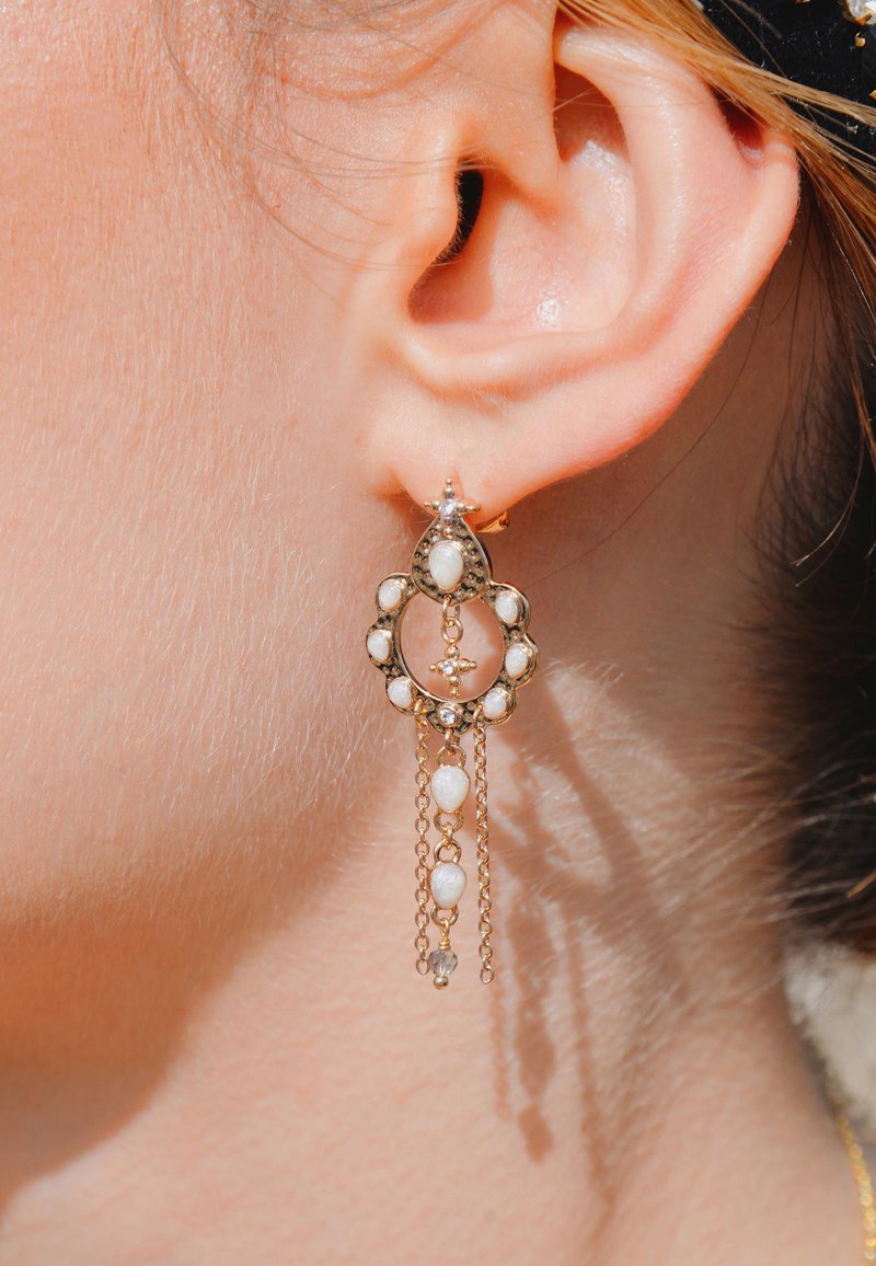 Les boucles d'oreilles pendantes en or ont un design orné avec un cadre circulaire, des accents en pierre blanche et plusieurs chaînes suspendues en dessous.