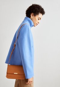 Jasnoniebieska bluza polarowa z wysokim kołnierzem, w połączeniu z brązową skórzaną torbą na ramię z zapięciem. Modelka ma na sobie złote kolczyki koła.