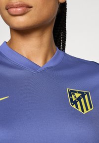 Maglia atletica viola a V con tessuto strutturato, caratterizzata da un logo Nike giallo e uno stemma con strisce e stelle blu e gialle.