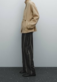 Beige liinake jakk nupuga esi ja vöödiga, koos mustade ja hallide vertikaalsete triibuliste pükstega. Mustad kingad täiendavad riietust.