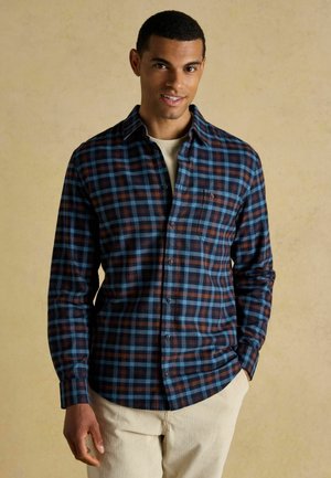 Button-down-Hemd aus Flanell im dunkelblauen, braunen und hellblauen Karomuster. Es hat eine Tasche und einen runden Saum. Kombiniert mit beigen Cordhosen.