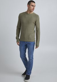 Olivgrüner Strickpullover mit strukturiertem Muster, Rundhalsausschnitt, kombiniert mit hellblauen Jeans und schwarzen Sneakers.
