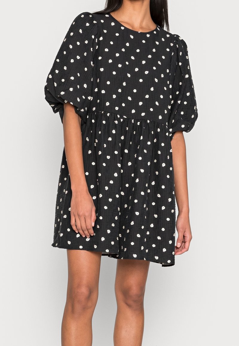 Mustapuku, jossa on valkoisia kukkakuoseja, lyhyet puffihihat, pyöreä pääntie ja levenevä siluetti. Rakennekangas lisää syvyyttä.