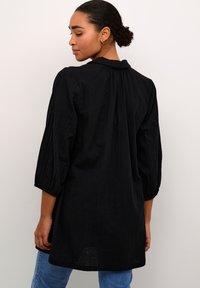 Tunique noire en tissu texturé, avec des manches trois quarts et un col montant. Coupe décontractée, l'ourlet tombe en dessous des hanches.