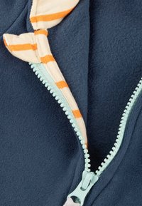 Marineblaues Fleece-Oberteil mit einem hellblauen Reißverschluss und gestreiftem Innenfutter in Creme und Orange. Der Reißverschluss hat einen strukturierten Zippfinger.