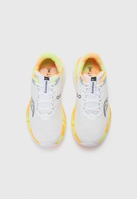 Zapatillas deportivas blancas con parte superior de malla, que presentan acentos en naranja y amarillo neón en la suela, y un logotipo negro en el lateral.