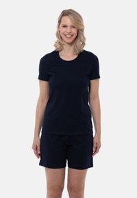 Leende kvinna med axellångt blont hår som bär en svart kortärmad tröja och svarta shorts, stående framför en enfärgad bakgrund.