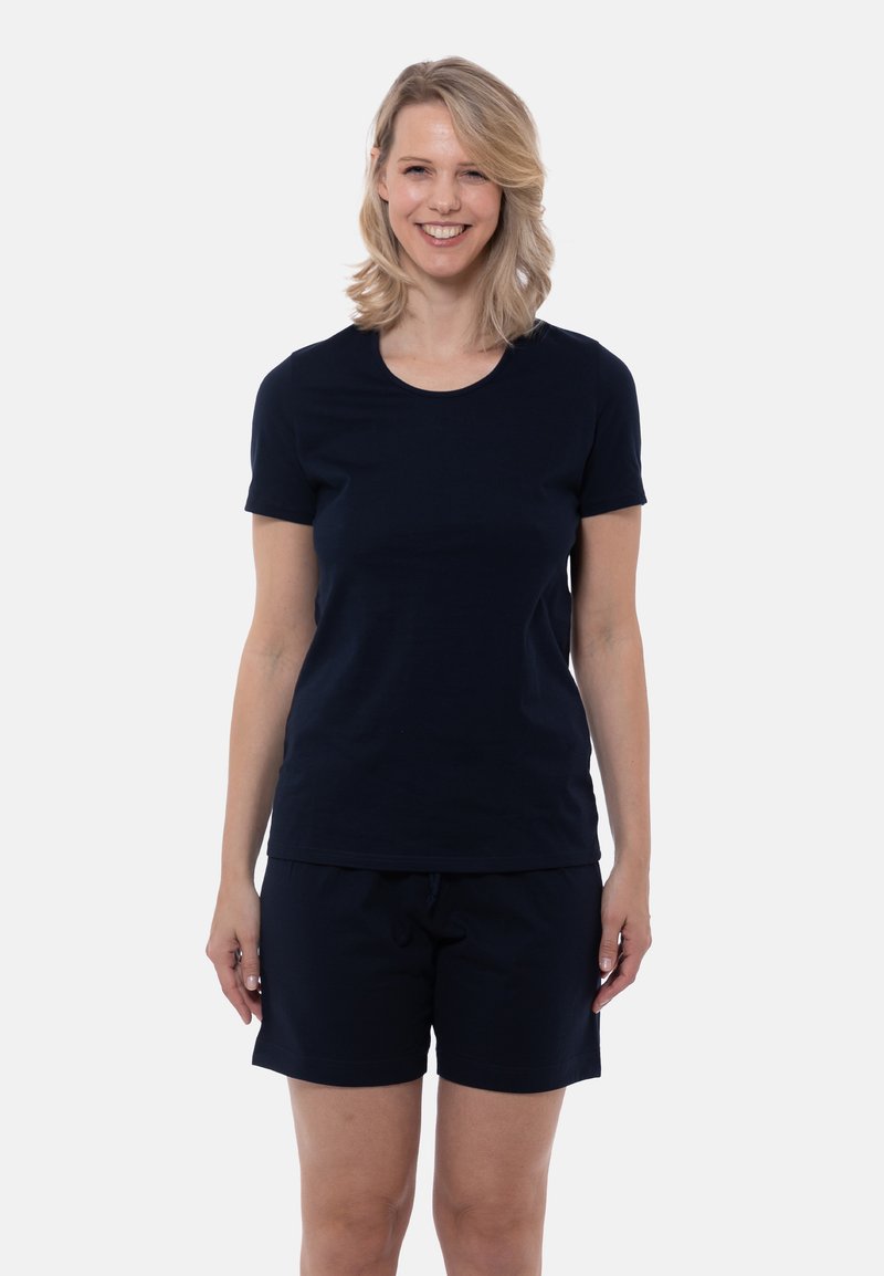 Leende kvinna med axellångt blont hår som bär en svart kortärmad tröja och svarta shorts, stående framför en enfärgad bakgrund.