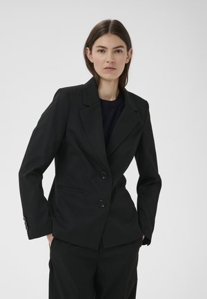Frau trägt ein tailliertes schwarzes Sakko mit zwei Knöpfen über einem dunklen Oberteil, steht mit den Händen in den Taschen vor einem schlichten Hintergrund.