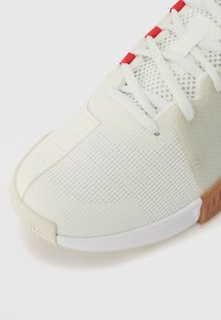 Chaussure de sport blanche avec tige en mesh, motif texturé, accents rouges près des lacets et semelle en caoutchouc. Comprend un orteil renforcé et un col rembourré.