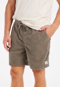 Brune kordfløjlshorts med snor i taljen, der har lodrette striber og et lille logo-patch på venstre ben. Glat tekstur.