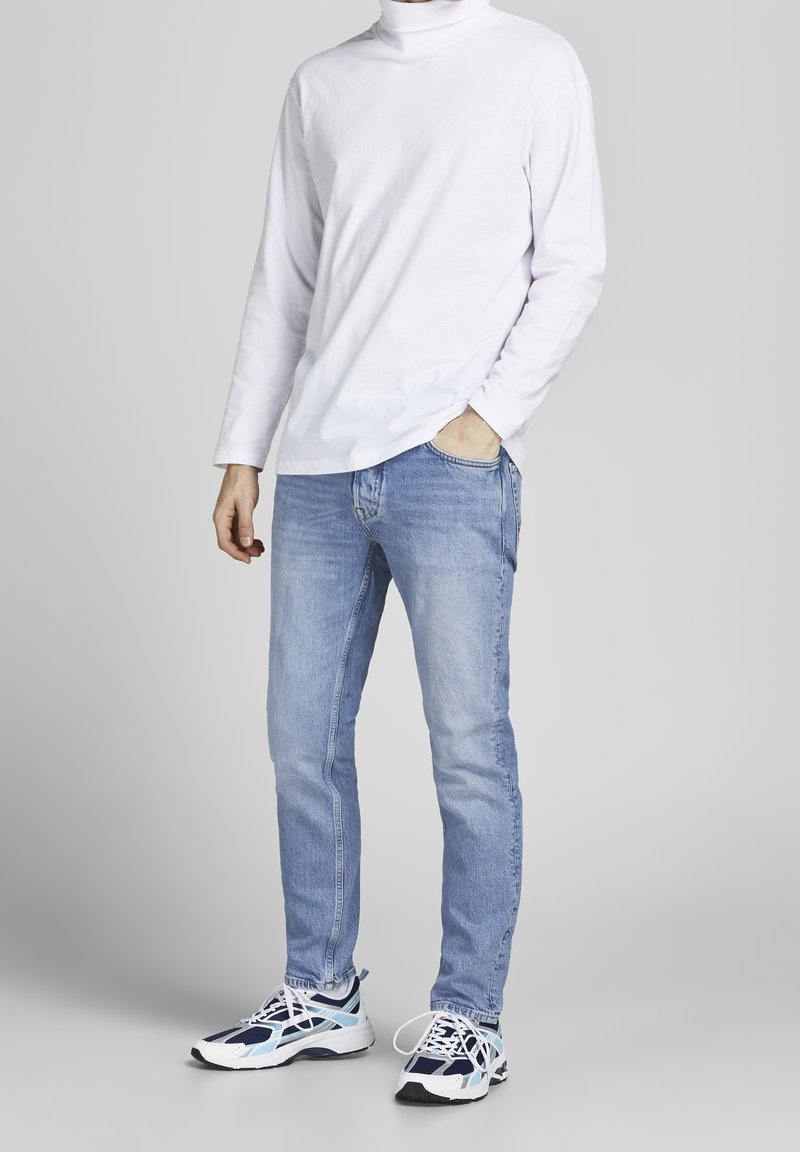 Un turtleneck blanc à manches longues, un jean bleu clair, et des chaussures de sport aux accents de blanc, marine et turquoise. Coupe décontractée.
