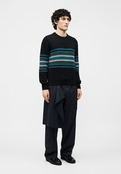 Jeune homme aux cheveux bouclés portant un pull noir avec des rayures bleues et grises, ainsi qu'un pantalon ample à fines rayures sombres avec un détail de tissu drapé.