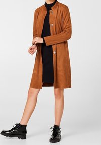 Manteau long en daim marron avec un col mandarin, deux poches avant et des accents de boutons en argent, porté sur une robe noire ajustée et des bottes noires.