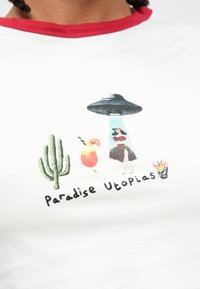 Άσπρο μπλουζάκι με κόκκινο γύρο λαιμού που απεικονίζει πολύχρωμο γραφικό: έναν κάκτο, κοκτέιλ, φιγούρα χούλα, UFO και το κείμενο "Παράδεισος Ουτοπίες."
