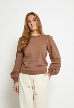 Kvinde med langt blondt hår iført en brun sweatshirt med ballonærmer og cremefarvede bukser med vide ben og stofbælte mod en hvid baggrund.