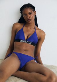 Modrý pruhovaný bikini s trojuholníkovým vrchom a logom na páske, spolu s zodpovedajúcimi bokovým viazaním. Hladká textúra s minimálnymi detailmi z kovu.