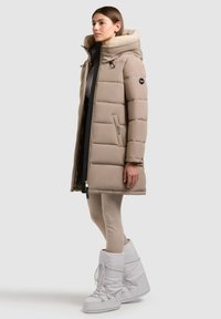 Junge Frau im Profil stehend, trägt einen beigefarbenen, gepolsterten langen Mantel mit fellgefütterter Kapuze, beige Leggings und weiße, gepolsterte Winterstiefel.