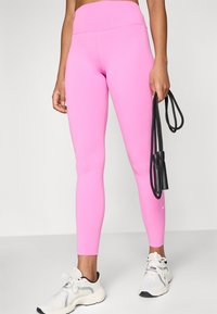 Rosa atletiska tights gjorda av stretchigt tyg med hög midja. Bärs med vita och svarta sneakers. Svart hopprep i handen.