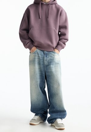 Lila Kapuzenpullover mit einer Fronttasche, kombiniert mit hellblauen weiten Jeans und grauen Sneakers mit weißen Akzenten.