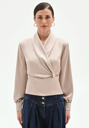 Donna con capelli scuri che indossa orecchini a cerchio dorati, una blusa beige a maniche lunghe avvolgente e jeans blu scuro plissettati a vita alta.