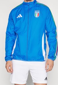 Blå lättviktsjacka med hel dragkedja, med vita och gröna detaljer. Framsidan visar Italiens logotyp och Adidas-logotyp på bröstet.