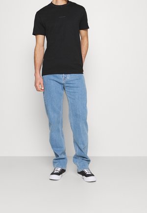 Muž v jednoduchém černém tričku s krátkým rukávem, světle modrých rovně střižených džínech a černobílých teniskách stojí na neutrálním pozadí.