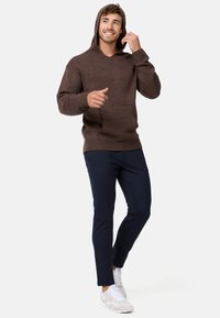 Haut en tricot marron avec un large col, des poignets côtelés et une poche kangourou, associé à un pantalon bleu marine ajusté et des baskets blanches.