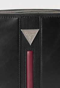 Μαύρο δερμάτινο πορτοφόλι με υφασμάτινη επιφάνεια, κόκκινη λωρίδα διακόσμησης και τριγωνικό ασημένιο λογότυπο με διακοσμητικό μοτίβο. Κλείνει με φερμουάρ.