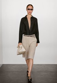 Modell klädd i en svart oversized skjorta med knapp, beige mockashorts, svarta plattformssandaler och håller en vit handväska.