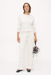 Kremfarget strikket genser med lange ermer, kombinert med hvite joggebukser med snor. Modellen holder brune høye hæler. Enkel og moderne design.