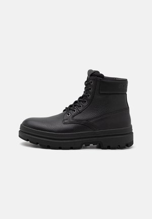 Zwarte leren enkellaars met gestructureerde afwerking, ronde neuzen, zes oogjes en een robuute zool. Heeft een gevoerde kraag en subtiele branding.