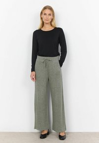 Schwarzes Langarmoberteil kombiniert mit weit geschnittenen, hellgrünen Strickhosen mit Kordelzugbund. Das Model trägt schwarze Schuhe.