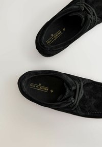 Mustat mokkakengät, joissa on pyöreä kärki, nauhat ja teksturoitu päällinen. Sisäpuolella on kultaiset kirjaimet mustalla taustalla.