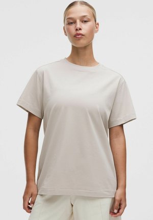 Ung kvinde med blondt hår trukket tilbage bærer en løs, lys beige kortærmet t-shirt og matchende lyse bukser mod en ensfarvet baggrund.