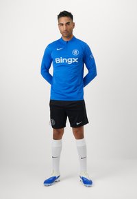 Blå langærmet sportstrøje med kvart lynlås og "Bingx" logo, parret med sorte shorts og hvide sokker, iført blå fodboldstøvler.