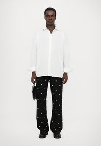 Cămașă albă cu nasturi, cu accente roșii, asociată cu pantaloni negri cu modele din puncte albe. Modelul ține o geantă mică neagră și poartă pantofi negri.