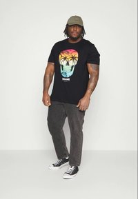 Svart T-skjorte med et fargerikt hodeskalle- og palmetre-grafikk, sammen med mørk grå jeans og svarte Converse-sneakers. Lys olivengrønn caps båret.