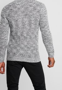 Mann in einem dicken, strukturierten grau-weißen Strickpullover mit langen Ärmeln und schwarzen Hosen, von hinten gezeigt.