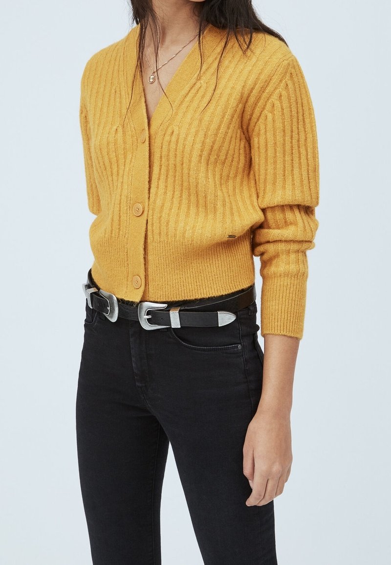 Kvinna klädd i en senapsgul ribbad kofta med knappar, svarta högmidjade jeans och svart bälte med silverdetaljer.