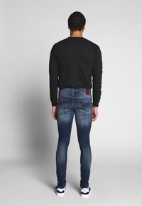 Camisa negra de manga larga, vaqueros ajustados azules con desvanecimiento, bolsillos y parche de cuero marrón. Las zapatillas blancas completan el atuendo.