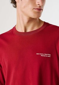 T-shirt rossa in cotone con collo rotondo e maniche corte. Sul petto è ricamato in bianco la scritta "Born to be a Rock Star; Forced to work."